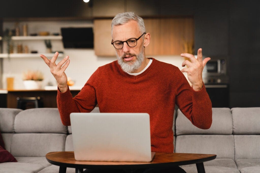 older man at laptop raising his arms as if frustrated
