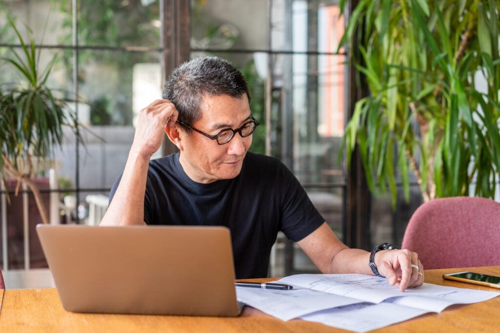older worker with laptop looking at documents and thinking