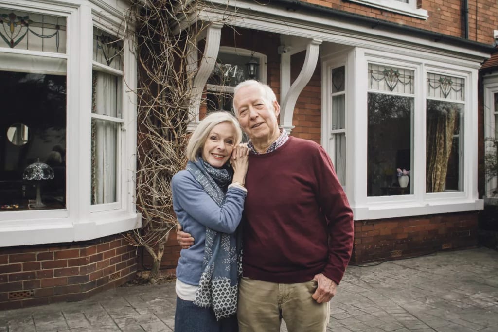 senior man and woman smiling and holding each other in front of a house home couple year mortgage.jpg
