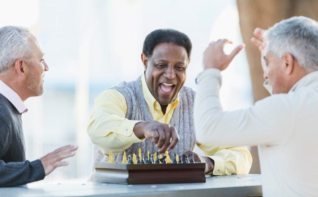 Three older people are sitting around a chessboard, laughing together.