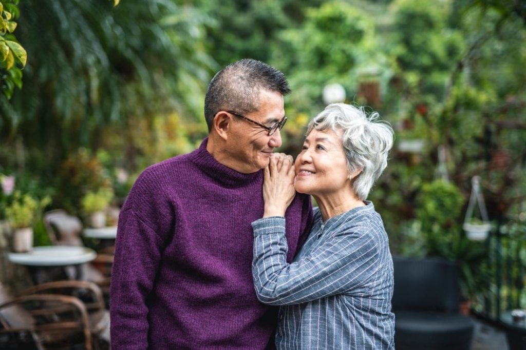Older couple standing in a leafy outdoor area, smiling at each other.