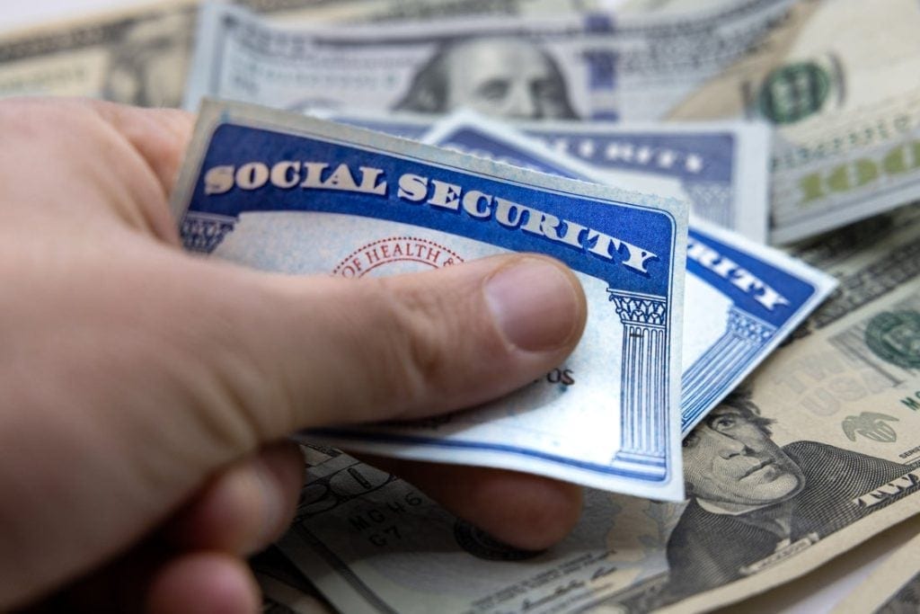 A hand holding two Social Security cards with a pile of American bills in the background.