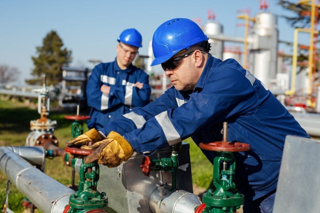 Two people standing in front of a pipeline with one turning a handle.