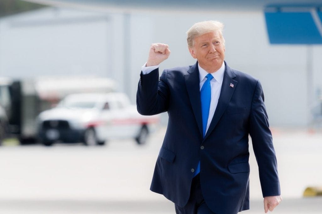 Donald Trump is raising his right hand in a fist while walking across an airport tarmac.