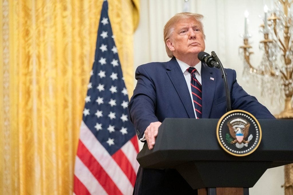 Donald Trump is standing behind a podium in the East Room of the White House.