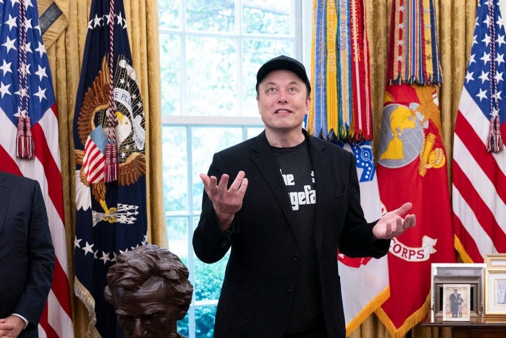 Elon Musk standing in the Oval Office with open hands and arms extended.