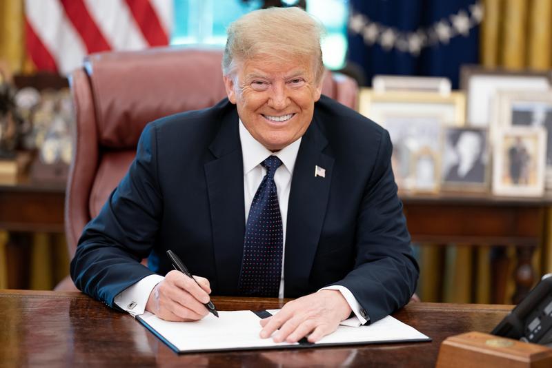 A smiling Donald Trump signing a bill in the Oval Office.